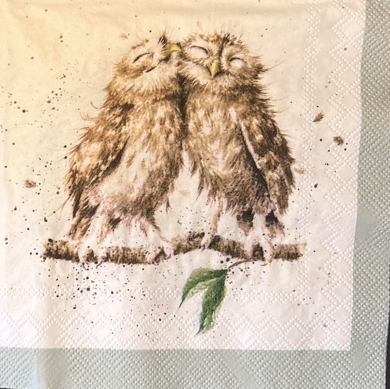 This picture of one of the napkins has two brown owls on a brown branch that has a green leaf, they have their heads side by side with each other with the appearance of bliss on their faces (etsy image id: 4852164401)