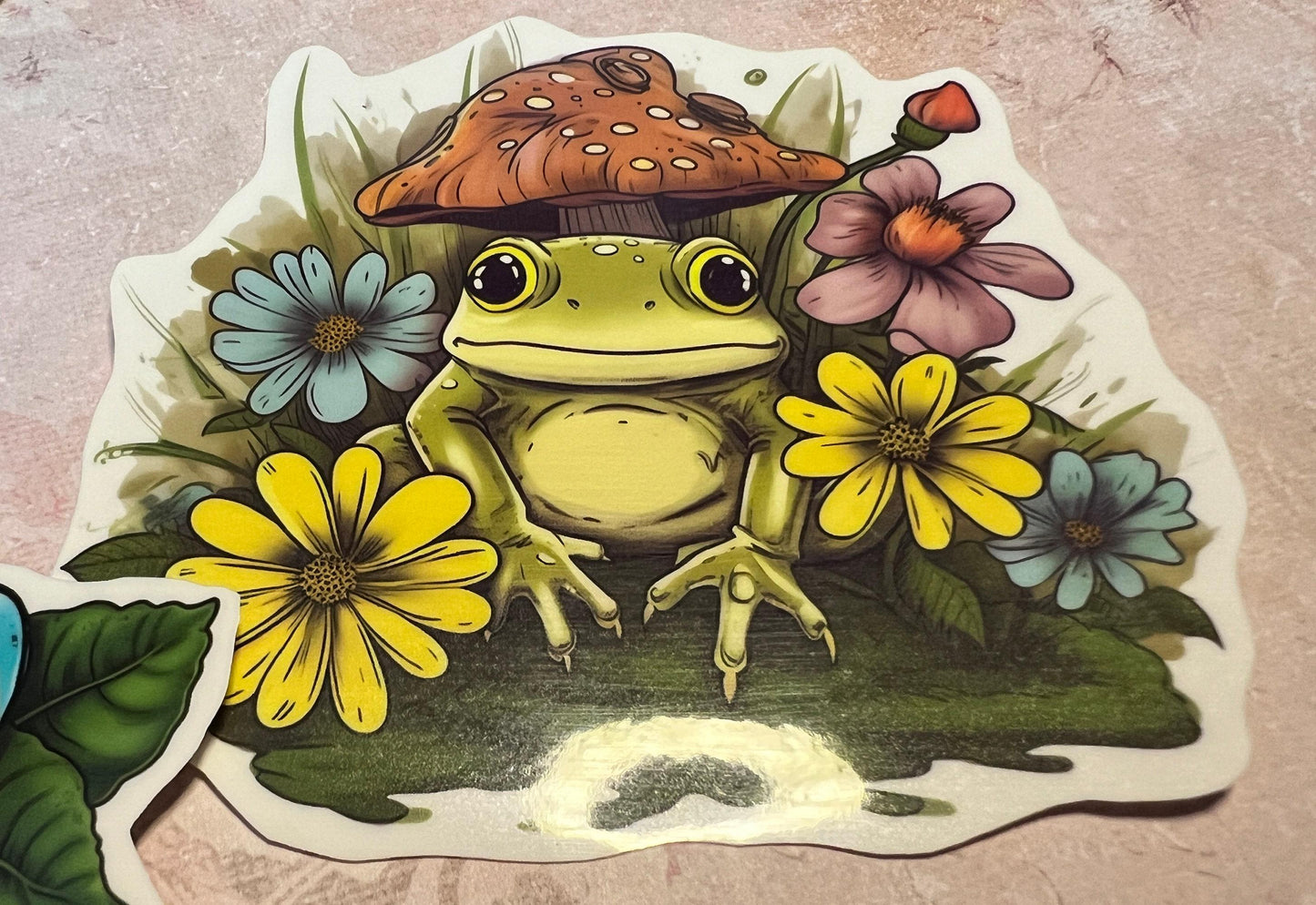 Green and yellow color Frog atop  greenery and flowers surrounding the image and a rust color mushroom in the background (etsy image id: 6694260797)